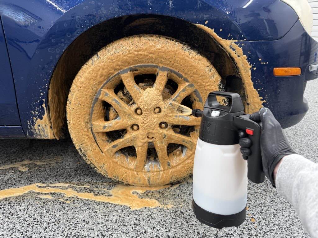 Dark Fury foaming tan across the alloy wheel and tire of a blue car, sprayed from a black hand pump sprayer at stronger than 4:1
