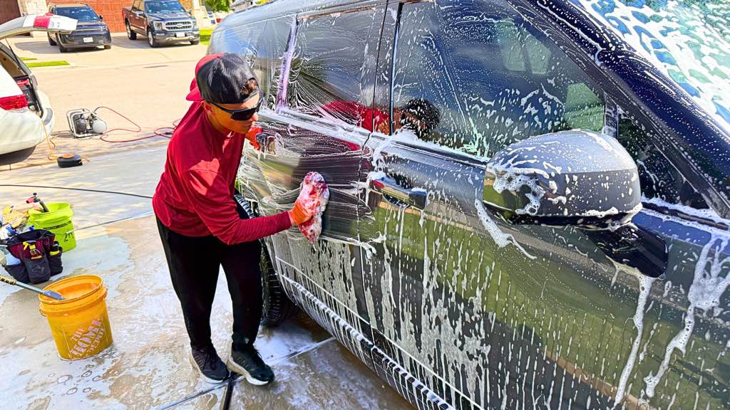 Detailer hand-washing a soapy black SUV with a wash mitt in direct sun, the paint still covered in car soap before the rinse